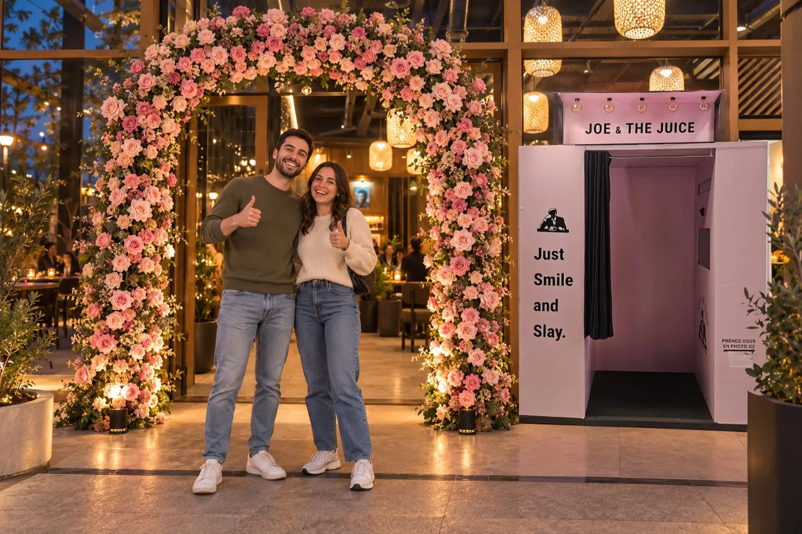 À l’entrée d’un café Joe & The Juice, un couple pose sous une arche de fleurs roses, à côté de la cabine photo rose « Just Smile and Slay »