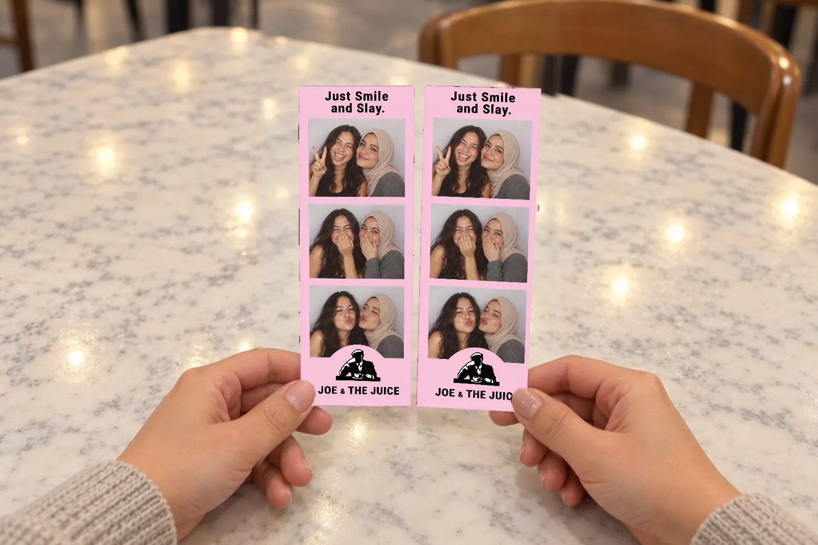 Deux amis prennent chacun une bandelette photo rose identique « Just Smile and Slay », imprimée en double, sur une table de café
