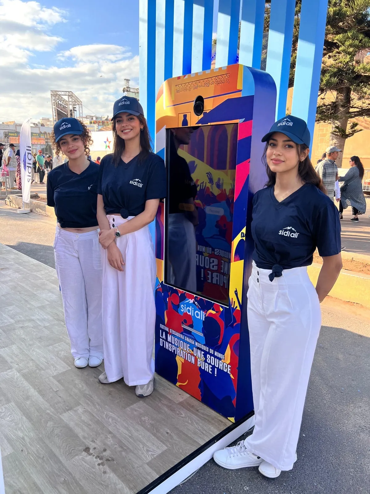 Trois hôtesses en tee-shirt et casquette Sidi Ali, souriantes, à côté de la borne habillée du slogan de la campagne
