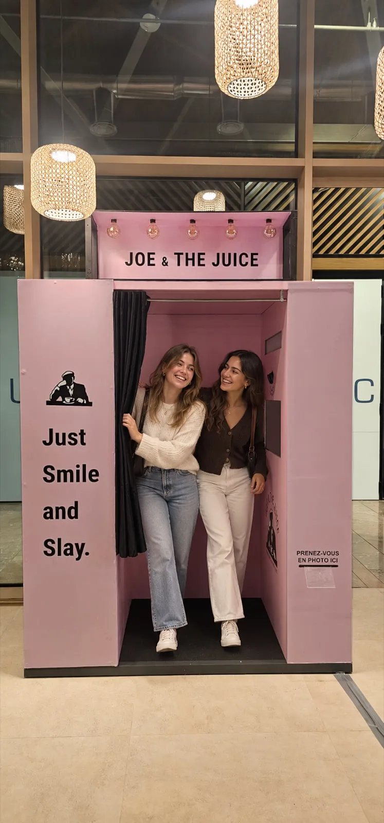 Deux amies souriantes sortent du rideau noir de la cabine photo rose Joe & The Juice « Just Smile and Slay », sous les suspensions en rotin du café