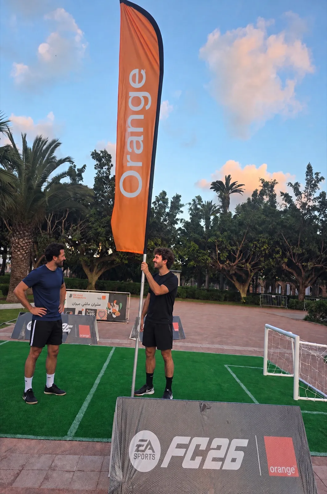 Un joueur dresse le fanion Orange au bord du mini-terrain habillé EA SPORTS FC 26, sous un ciel de fin de journée