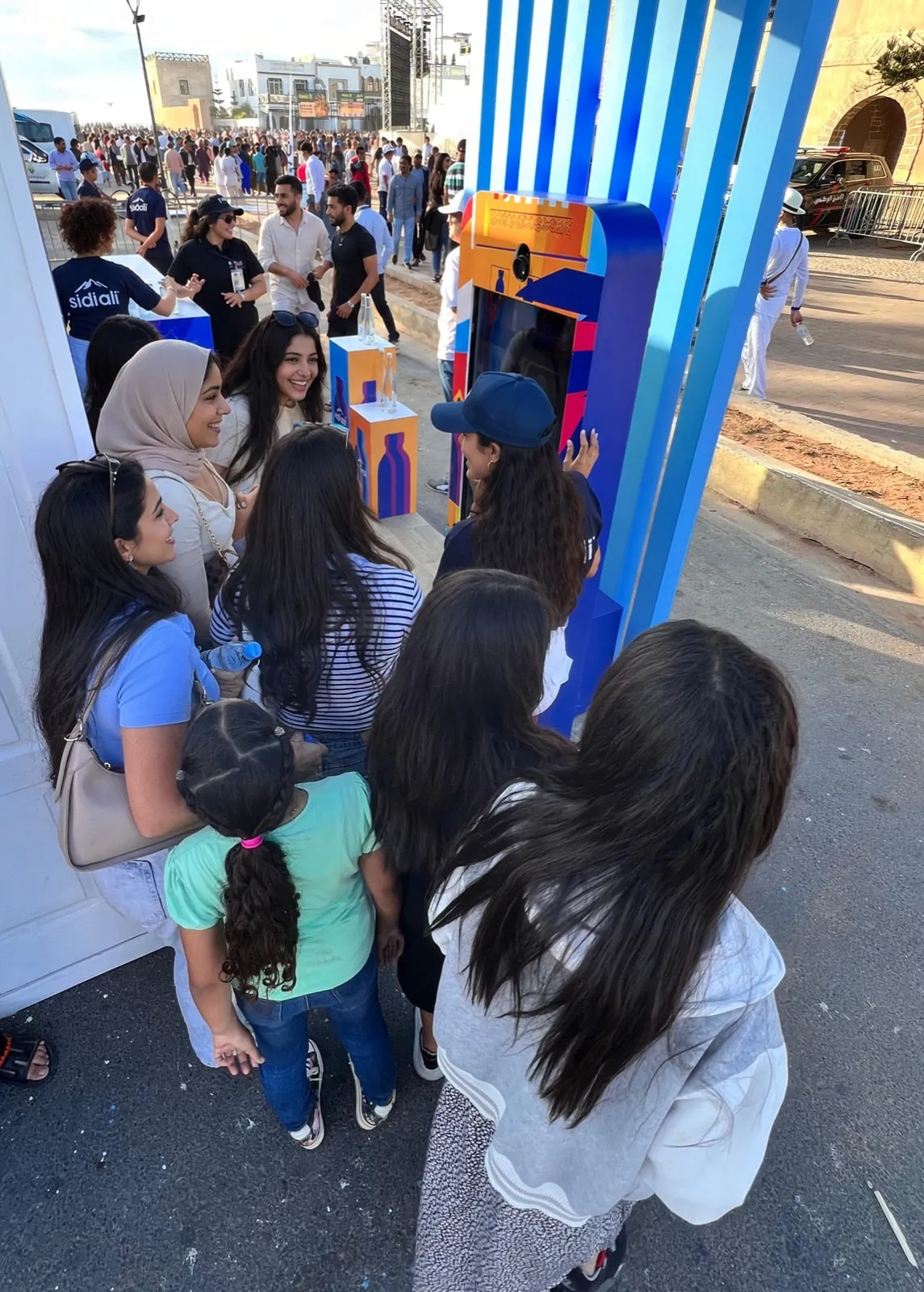 Un groupe de festivalières se presse autour de la borne Sidi Ali, une hôtesse les guide à l’écran, sur la promenade du festival