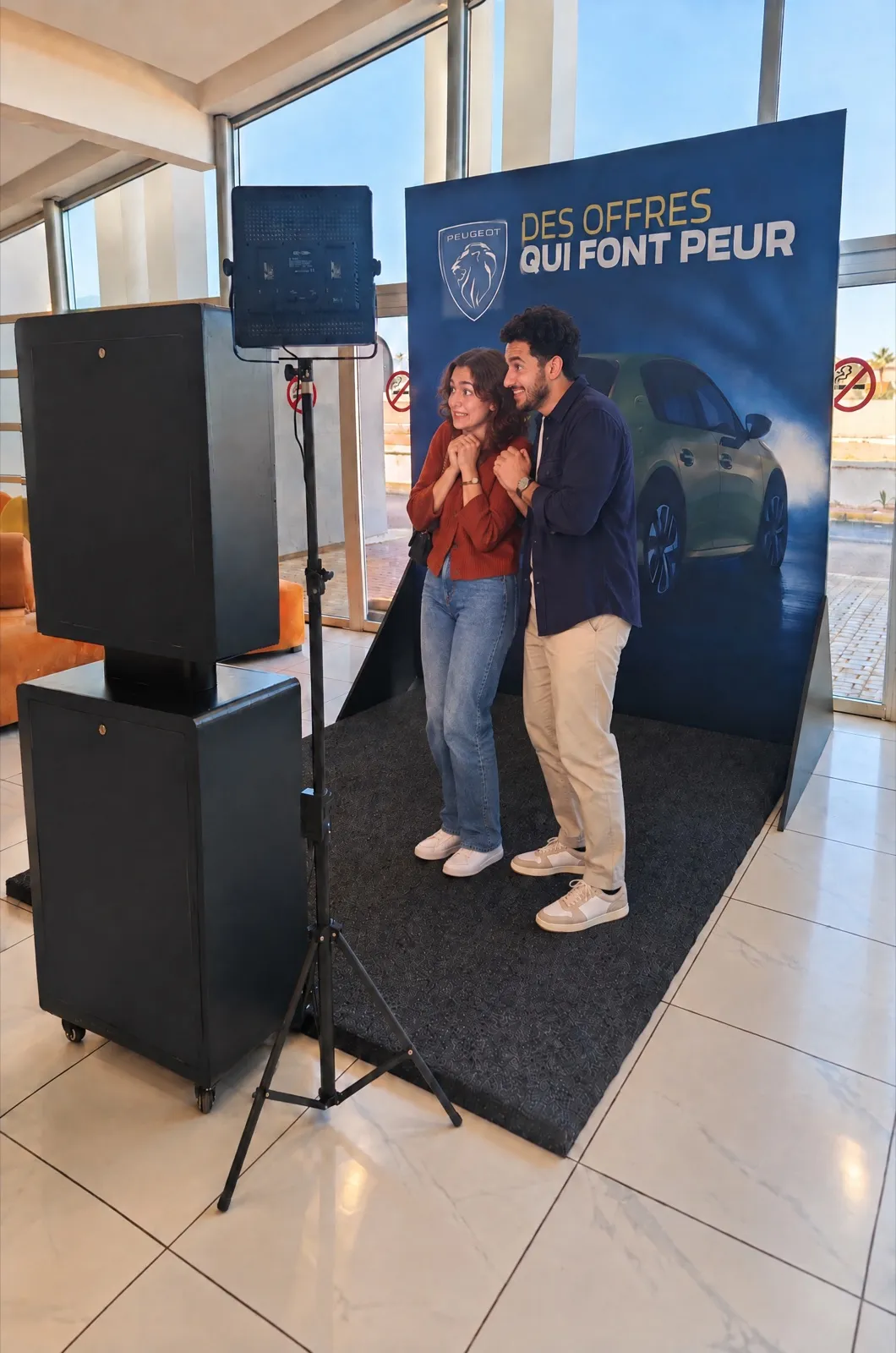 Devant le décor Peugeot « Des offres qui font peur », deux amis font mine d’avoir peur face à la borne photo