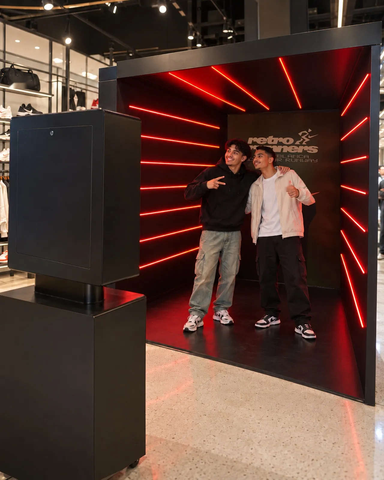 Deux amis posent dans le tunnel de néons rouges Retro Runners d’un magasin Nike, face à la borne photo noire habillée de la campagne