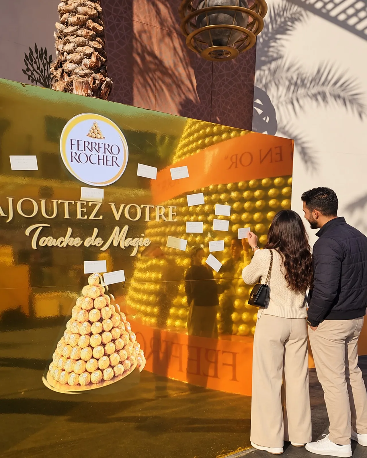 Un couple accroche sa carte de vœux sur le mur doré « Ajoutez votre touche de magie » de Ferrero Rocher