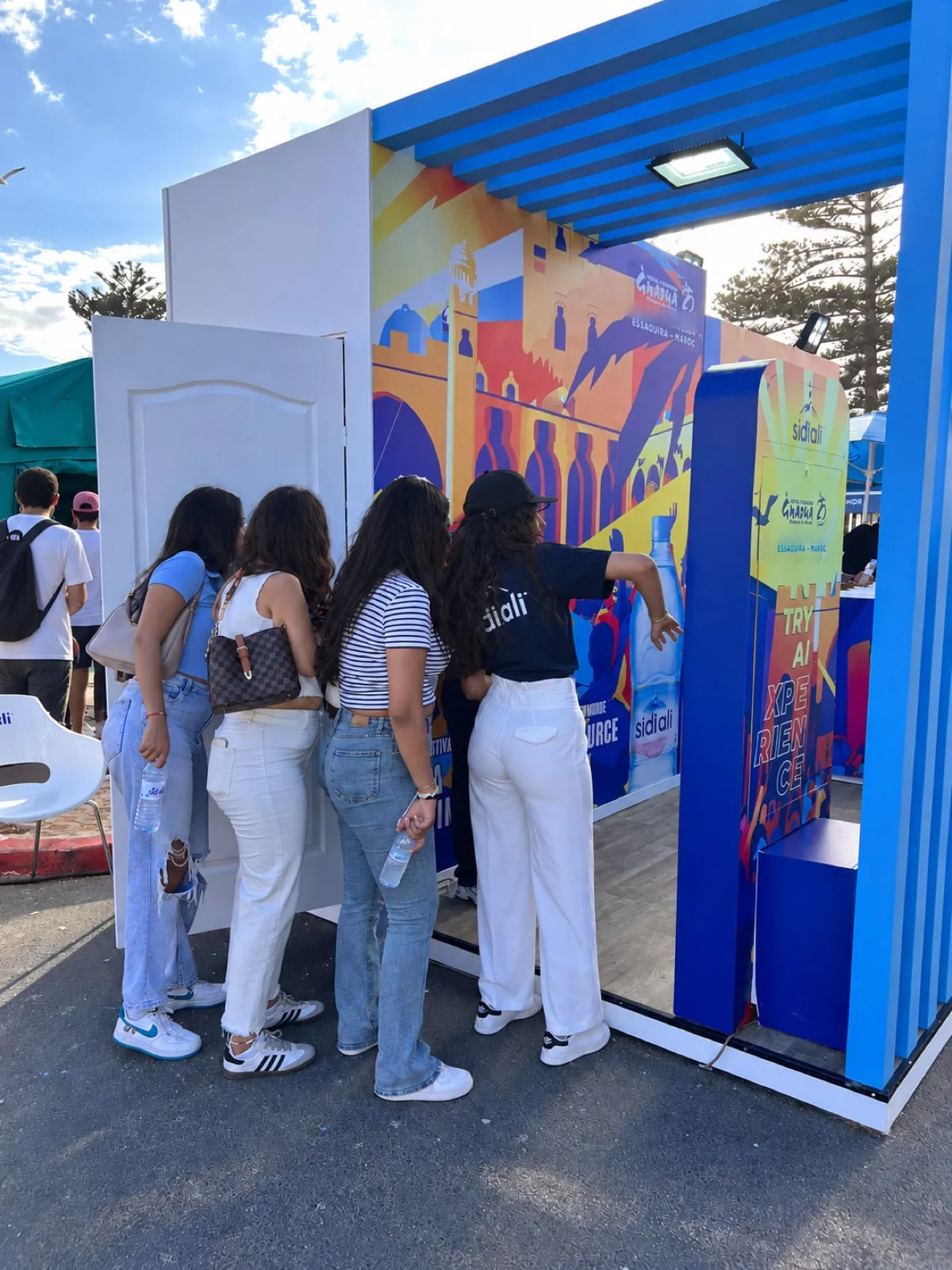 Un groupe d’amies se penche vers la borne pour découvrir son portrait, sous l’auvent bleu du stand