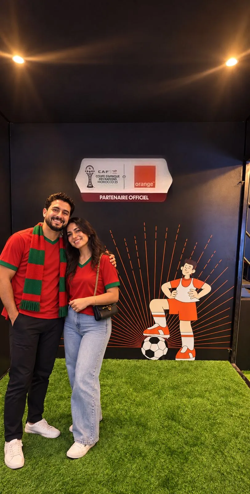 Un couple de supporters en maillot et écharpe du Maroc pose devant le photocall noir Orange, partenaire officiel de la CAN