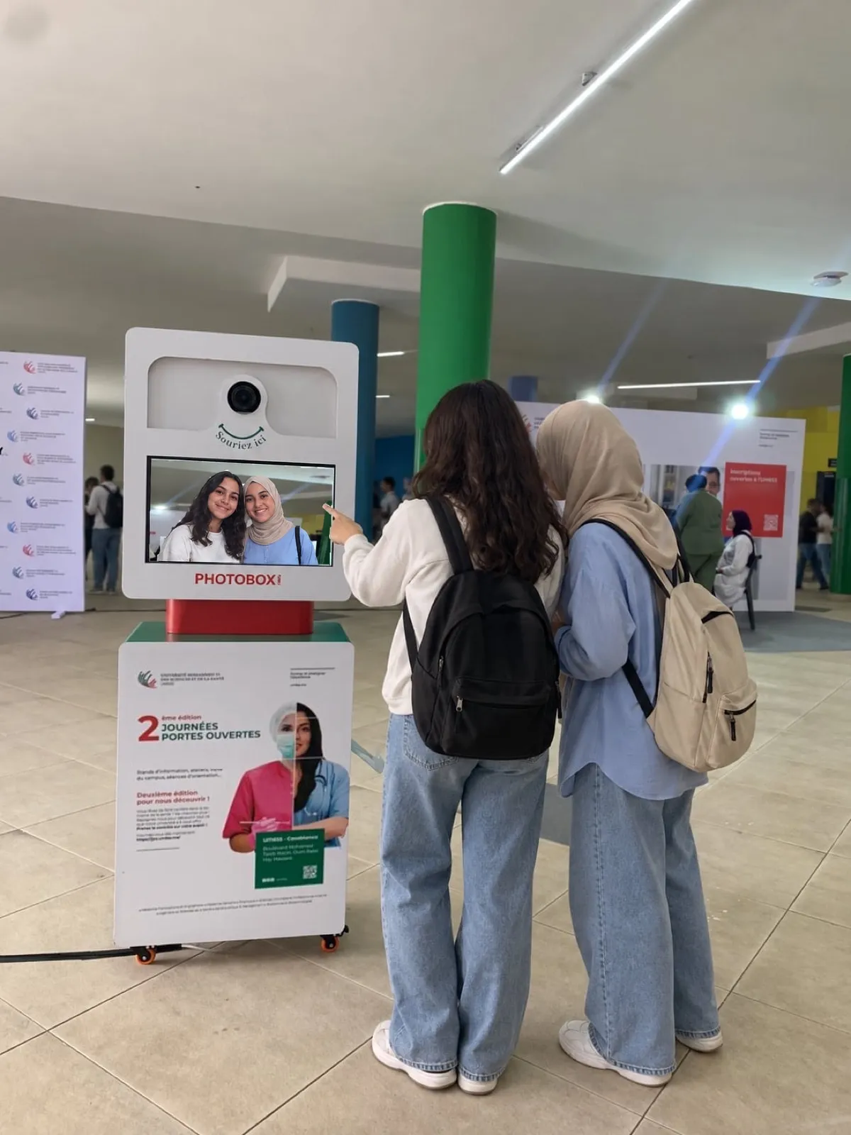 Deux visiteuses, de dos, découvrent leur photo sur l’écran de la Selfie Box habillée de l’affiche des portes ouvertes