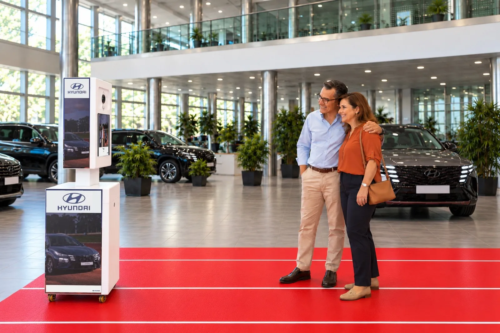 Vue du showroom Hyundai : de profil, la borne photo vise un couple qui pose sur le tapis rouge, le Tucson derrière eux