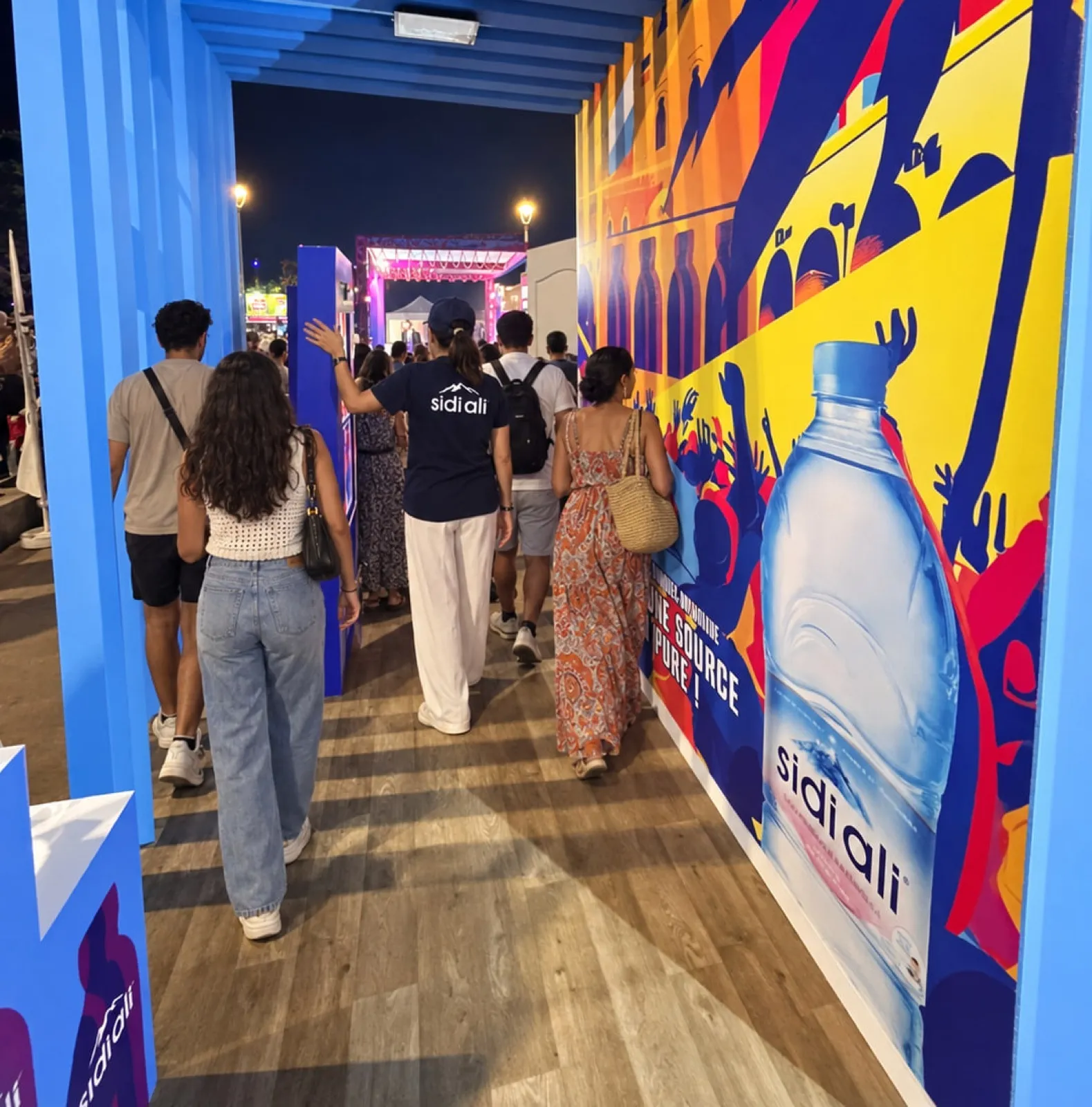 Des festivaliers avancent de nuit dans le couloir du stand Sidi Ali, entre une fresque de concert et une bouteille géante, au Festival Gnaoua