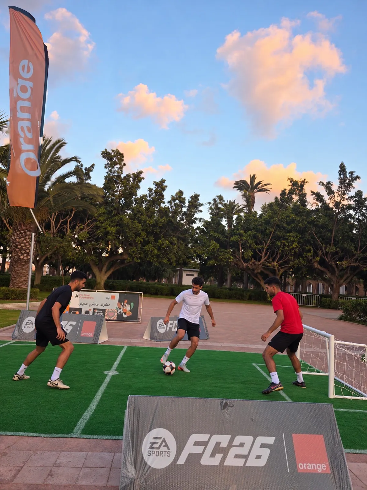 Le mini-terrain habillé EA SPORTS FC 26 et Orange, fanion dressé, où de jeunes joueurs échangent quelques passes en fin de journée