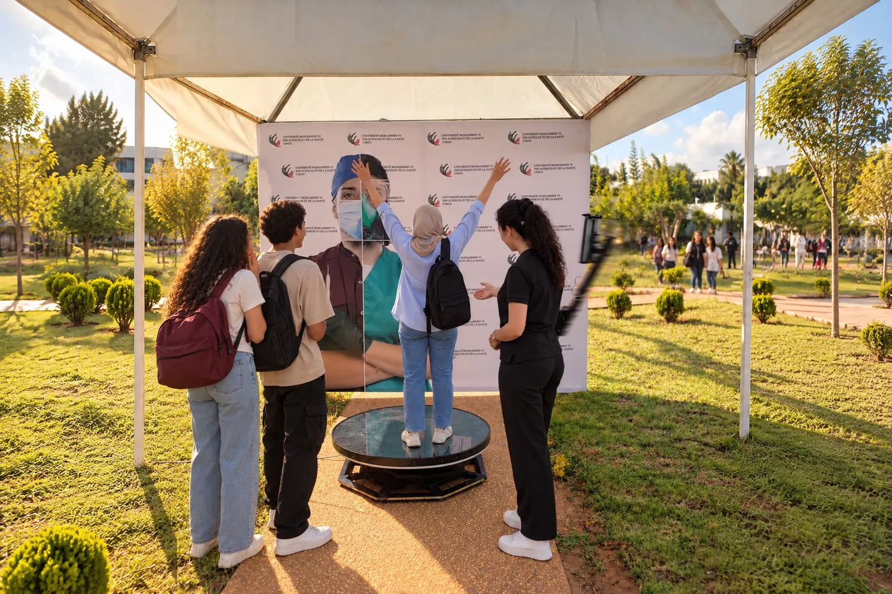 Sous une tente du campus, une visiteuse lève les bras sur le plateau de la borne 360°, guidée par une hôtesse PhotoBox ; deux amis attendent