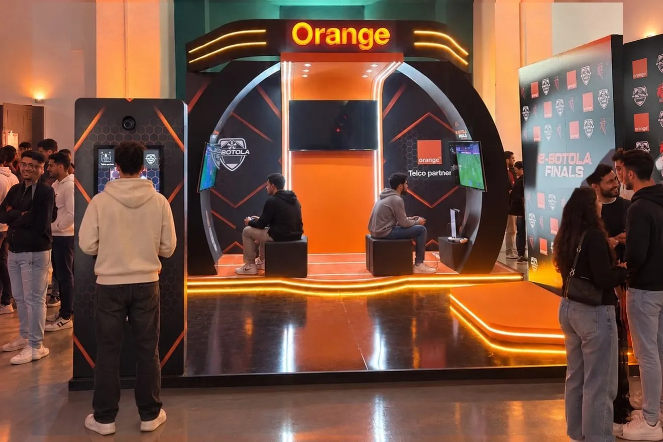 Le stand Orange des e-Botola Finals sous son arche lumineuse : deux joueurs s’affrontent sur EA SPORTS FC 26, un visiteur devant la borne PhotoBox