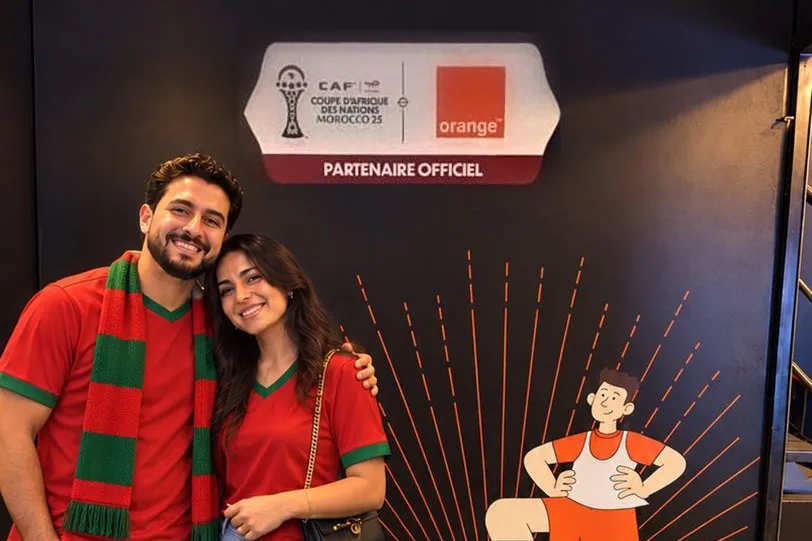 Un couple de supporters en maillot du Maroc, écharpe au cou, sourit devant le photocall noir Orange, partenaire officiel de la CAN