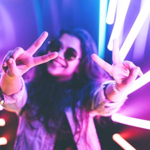 Cheerful cute smiling girl shows peace sign in round glasses at purple neon background.